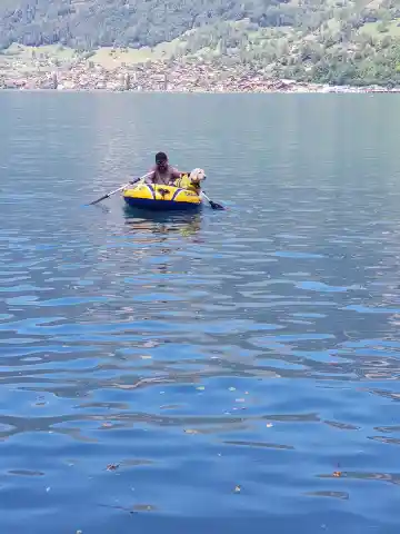 Eine Person sitzt mit einem weissen Hund in einem gelben Schlauchboot auf einem See vor bewaldeten Berghängen.