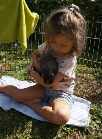 Ein kleines Mädchen sitzt auf einer Decke im Gras vor einem Gitterzaun und hält einen dunklen Welpen im Arm.
