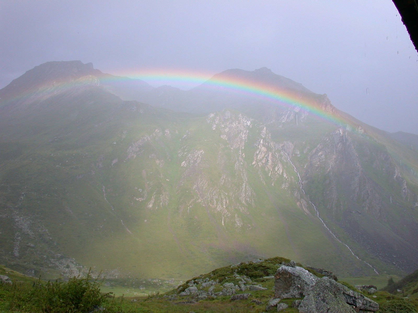 Ein Regenbogen spannt sich über einen grünen, felsdurchsetzten Alphang im Regen, in den Runsen fallen Wasserfälle.